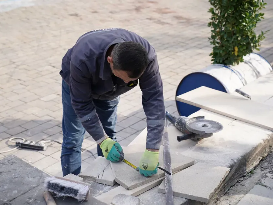 Stratenmaker in werkkleding meet een betonnen tegel af voor het zagen, met een haakse slijper ernaast